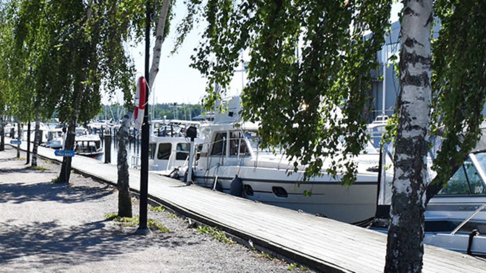 Längs med kajen och bryggan från Tullhuset till gästhamnen ligger större och mindre båtar med långsidan mot kajen. Promenaden bredvid bryggan är omgiven av björkar och träd.