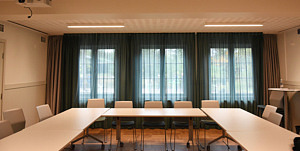 Bord och stolar står uppradade, i bakgrunden skyms tre stora fönster av skira gardiner i blått