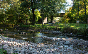 Nere vid parkmiljön i Husby park ligger stenar utmed åkanterna och vattnet ringlar sig förbi. I bakgrunden syns parkmöbler under höga träd. Solen strilar genom lövverket.