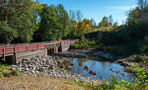Ovan bron vid Husby kvarn efter fiskepassageprojektet syns vattnet sakta rinna ner under gångbron. Stenar ligger utmed åkanterna och leder vattnet in genom två fåror under gångbron.