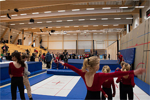 I förgrunden syns ryggen på unga gymnaster, framför dem syns de stora trampolinerna. I bakgrunden skymtar läktarna i trä.