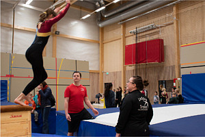 Flicka syns hoppa från plint mot en stor fluffig matta, två ledare tittar på gymnasten och står på varsin sida för att assistera.