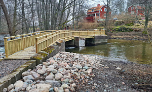 Ovan bron vid Nygårdsdammen efter fiskepassageprojektet syns högvattnet forsa under gångbron. Stenar ligger utmed åkanterna.