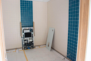 Helkaklad badrum i vitt och turkos där ett fäste för toalettstol syns på väggen.