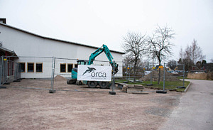 Uteplatsen vid Kulturskolans entré är inhägnad, en grön grävmaskin syns innanför och på stängslet syns en banderoll med Orcas logotyp.