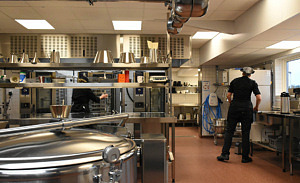 Ett stort kokkärl i förgrunden, en större arbetsbänk i bakgrunden och kockar som jobbar.