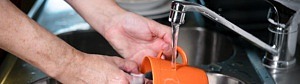 En närbild av en rostfri diskbänk där en persons armar skymtar, när de sköljer en orange mugg under kranens rinnande vatten.