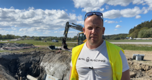 En man står framför utomhus med en grävskopa i bakgrunden.