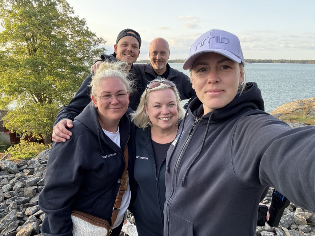 Gruppbild på fem leende personer. I bakgrunden syns träd och hav.