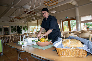 En person står vid ett bord med bröd, smör och bakom det bord och stolar.